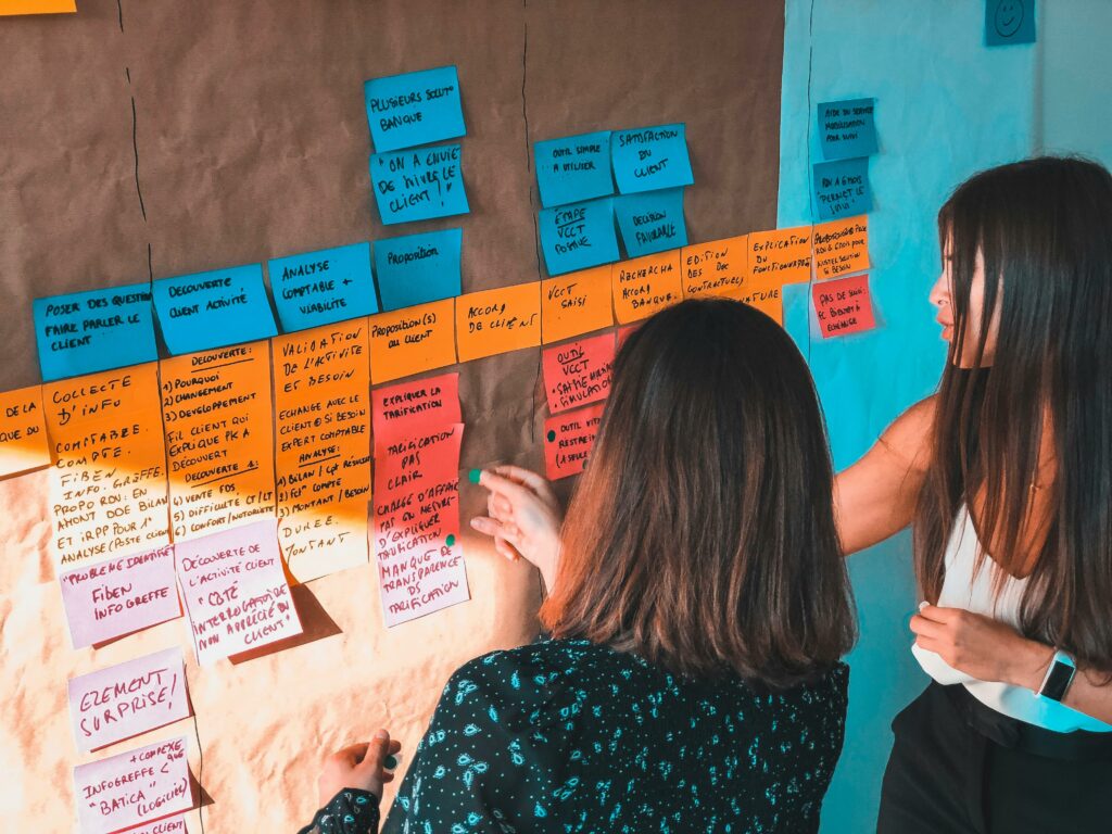 Two women engaged in collaborative planning using colorful Post-it notes on a board in a modern office space.